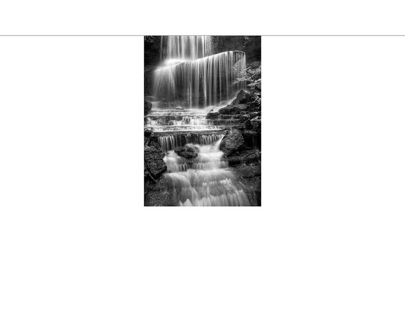 Ein beeindruckender Wasserfall fließt in Kaskaden über felsige Stufen, aufgenommen in einem stimmungsvollen Schwarz-Weiß-Stil.