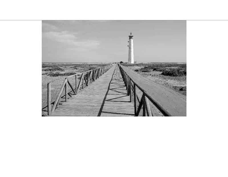 Schwarz-Weiß-Foto eines langen Holzstegs, der zu einem Leuchtturm führt, umgeben von einer weiten Landschaft