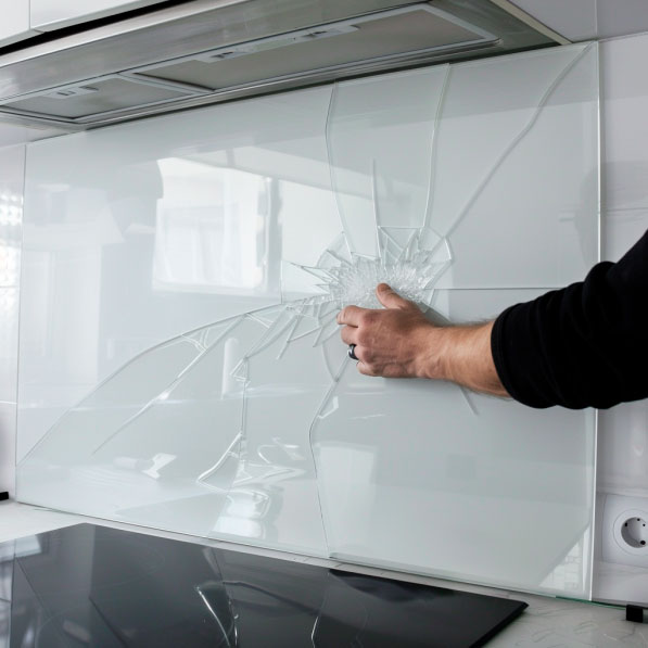 Nahaufnahme einer gebrochenen Glaswand in einer modernen Küche, berührt von einer Hand. Die Risse breiten sich sternförmig aus und sind deutlich sichtbar.