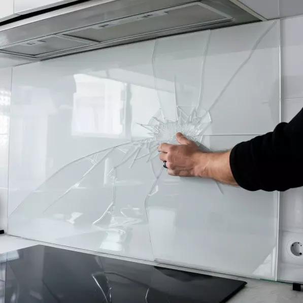Nahaufnahme einer gebrochenen Glaswand in einer modernen Küche, berührt von einer Hand. Die Risse breiten sich sternförmig aus und sind deutlich sichtbar.
