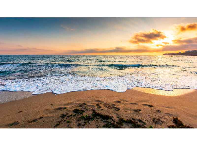 Ein Sonnenuntergang über dem Meer, die Wellen rollen an einen ruhigen Sandstrand.