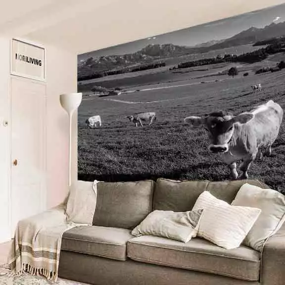 Ein Wohnzimmer mit einem großen Wandbild von Kühen auf einer Wiese vor Bergkulisse, einem beigen Sofa und Kissen.