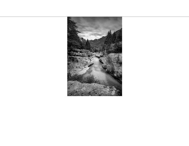 Ein ruhiger Fluss fließt durch eine felsige Schlucht, umgeben von Bäumen und Bergen im Hintergrund in einer Schwarz-Weiß-Aufnahme.