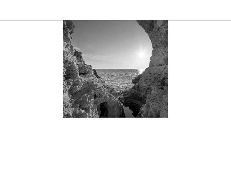 Steinklippen mit Meerblick bei Sonnenuntergang Ein schwarz-weißes Foto zeigt eine Öffnung zwischen Steinklippen, die den Blick auf das Meer und die untergehende Sonne freigibt.