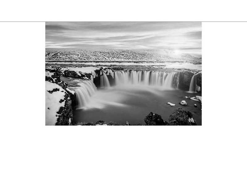 Ein Wasserfall fließt in einer winterlichen, schneebedeckten Landschaft mit ruhigem Himmel im Hintergrund.