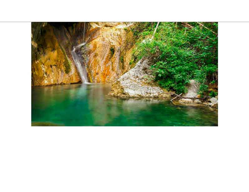 Ein idyllischer Wasserfall fließt in einen türkisfarbenen Teich, umgeben von üppigem Grün und Felsen im Wald.