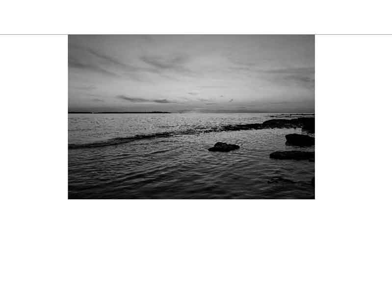 Schwarz-Weiß-Fotografie eines ruhigen Meeres mit Felsen im Vordergrund und bewölktem Himmel bei Sonnenuntergang