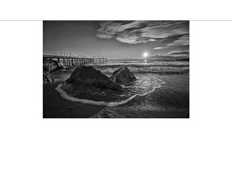 Schwarz-Weiß-Strandlandschaft mit Pier und Sonnenuntergang Eine dramatische Schwarz-Weiß-Aufnahme eines Strandes bei Sonnenuntergang mit einem Pier im Hintergrund und Wellen, die gegen Felsen branden.