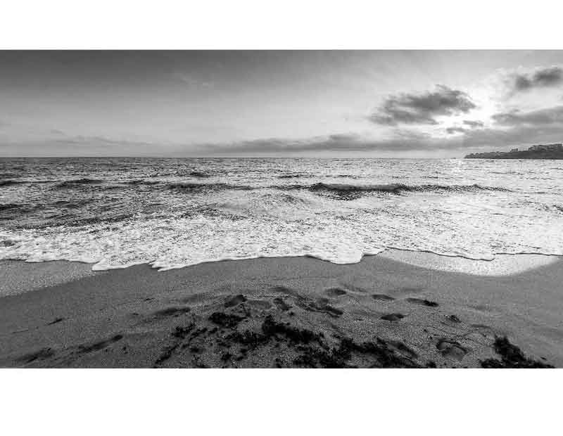 Ein schwarz-weißes Bild zeigt einen Strand mit Wellen, darüber ein bewölkter Himmel bei Sonnenuntergang.