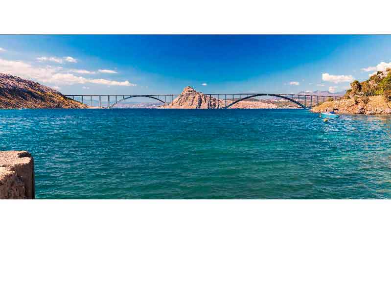 Panoramablick auf eine Brücke über türkises Wasser Eine große Brücke überspannt das weite türkisfarbene Wasser unter einem klaren blauen Himmel. Im Hintergrund sind Berge und eine hügelige Landschaft zu sehen.