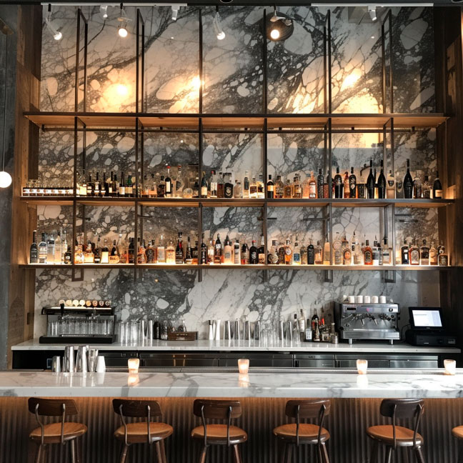 Innenansicht einer stilvollen Bar mit Marmorrückwand, hohen Regalen mit verschiedenen Spirituosenflaschen und gemütlichen Barhockern.
