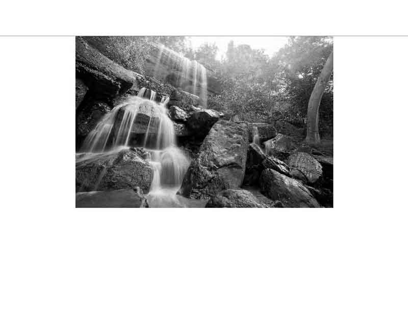 Ein malerischer Wasserfall fließt über Felsen in einem bewaldeten Gebiet, eingefangen in Schwarz-Weiß.