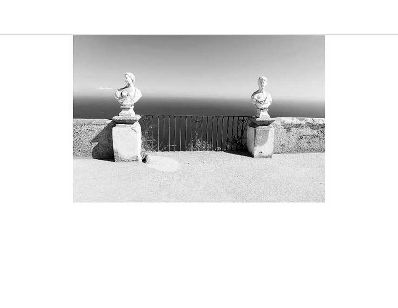 Zwei Steinbüsten stehen auf einer Terrasse mit Blick auf das Meer, eingefasst von einem Geländer.