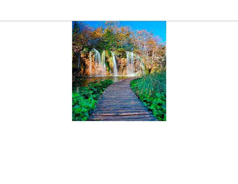 Ein Holzsteg führt durch grüne Vegetation zu beeindruckenden Wasserfällen, umgeben von herbstlichen Bäumen unter klarem Himmel.