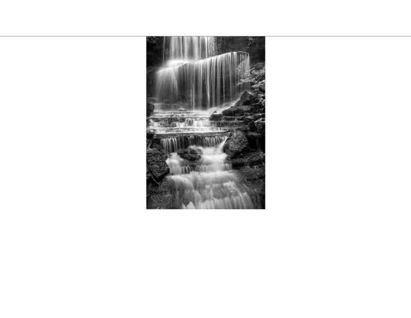 Ein Wasserfall fällt über Felsen, der Fluss fließt sanft durch den Wald im Schwarz-Weiß-Foto.