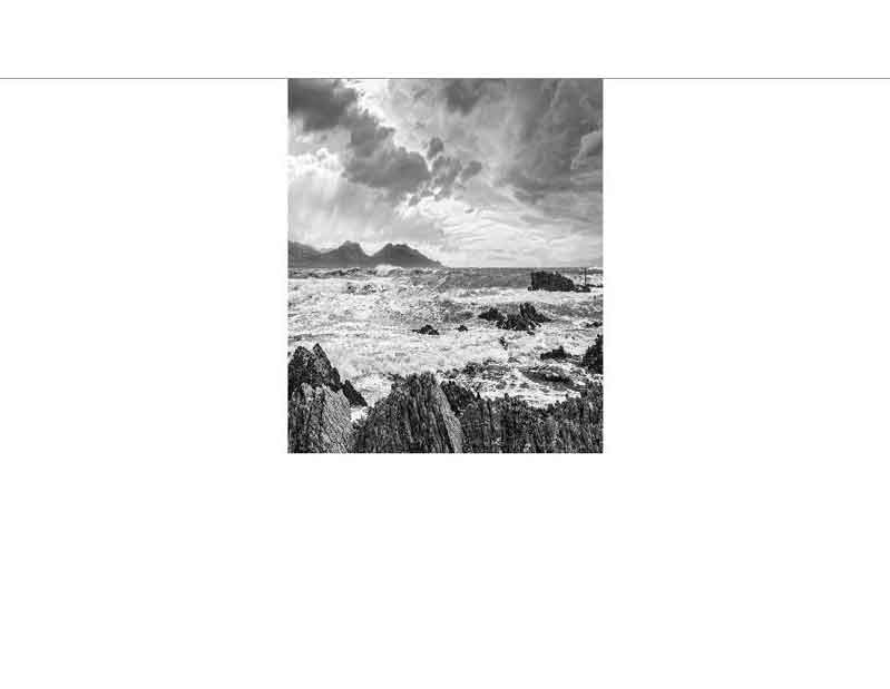 Schwarz-Weiß-Fotografie einer stürmischen Küste mit dramatischen Wolken und Felsen im Vordergrund.