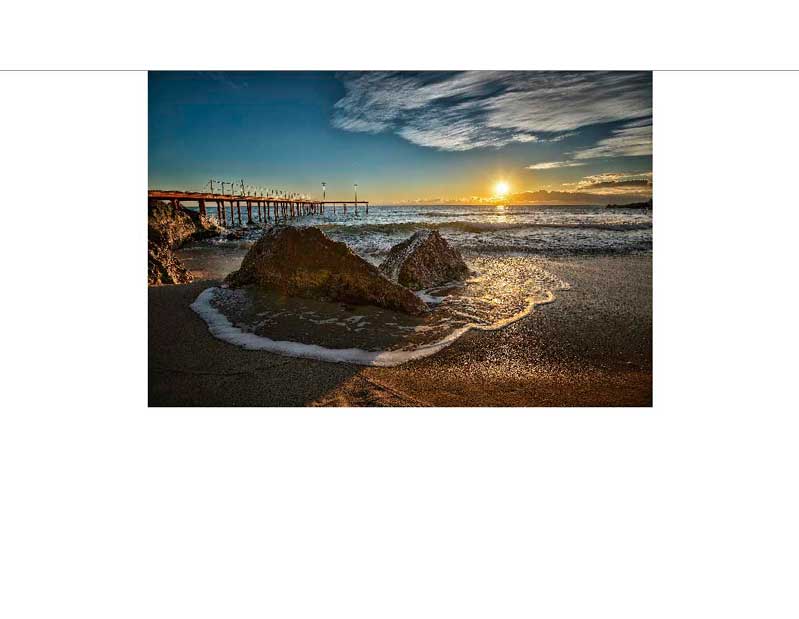 Idyllische Szene eines Sonnenuntergangs über dem Meer mit einem Steg und Felsen am Strand