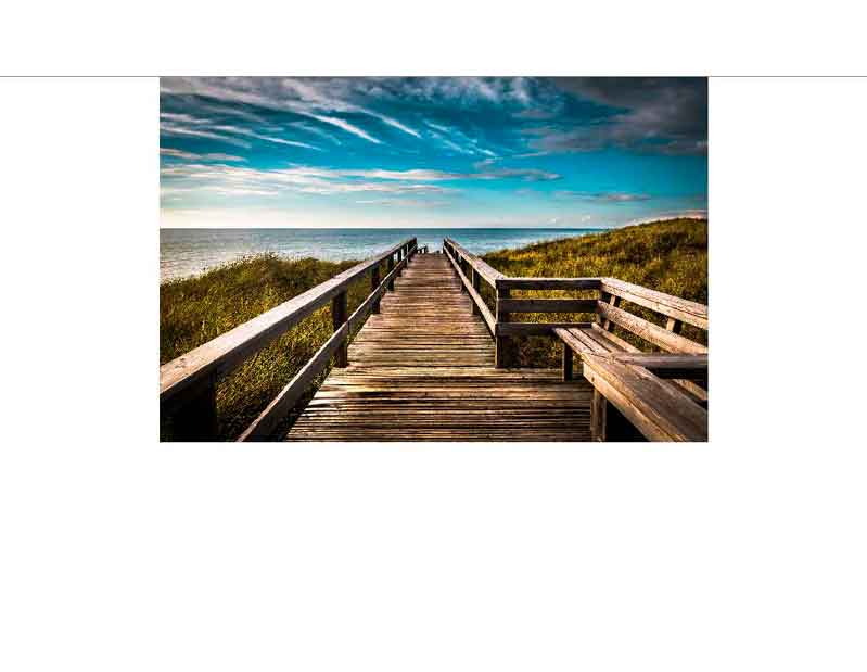Ein langer Holzsteg führt durch Dünen zum Meer, mit blauem Himmel und wenigen Wolken