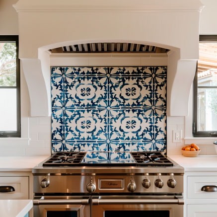 Elegante Küche mit einem großen Edelstahlherd und blauen, kunstvoll gemusterten Fliesen als Rückwand, flankiert von weißen Wänden und Naturlicht durch Fenstern links und rechts.