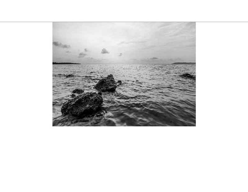 Schwarz-Weiß-Fotografie eines ruhigen Meeres mit Felsen Schwarz-Weiß-Bild eines ruhigen Meeres mit großen Felsen im Vordergrund bei bewölktem Himmel.