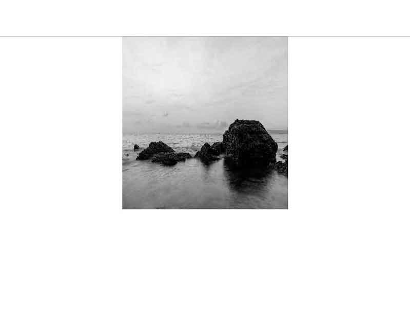 Ein ruhiger Küstenabschnitt mit großen Felsen im Wasser, aufgenommen in Schwarz-Weiß.