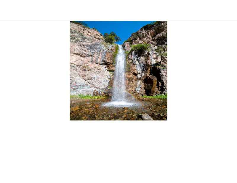 Ein hoher Wasserfall, der über eine felsige Klippe in ein klares Wasserbecken stürzt, umgeben von grüner Vegetation unter einem strahlend blauen Himmel.