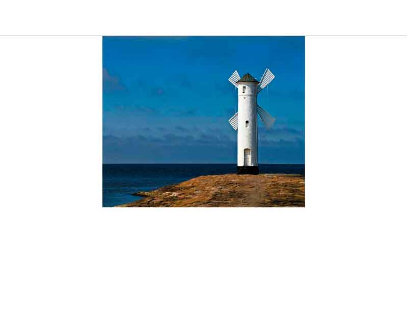 Ein weißer Leuchtturm mit Windmühlenflügeln an der Küste unter blauem Himmel