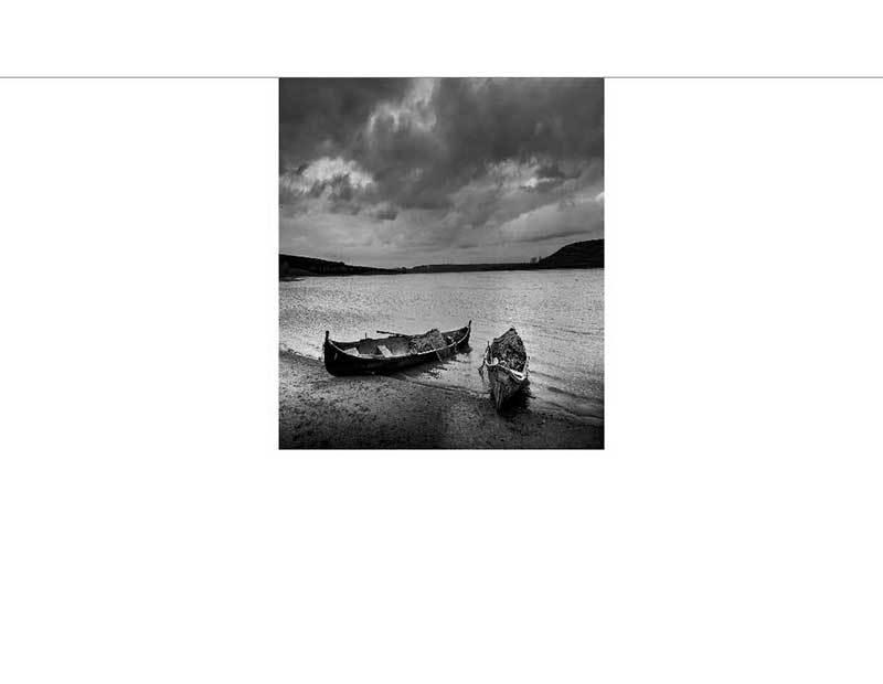 Schwarz-Weiß-Foto von zwei Holzbooten am Seeufer unter einem wolkenverhangenen Himmel