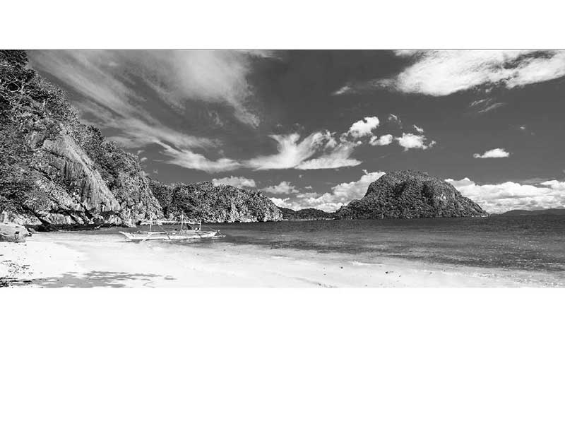 Schwarzweißfoto eines Strands mit einem Boot und Felsen im Hintergrund, ruhiges Meer und bewölkter Himmel.