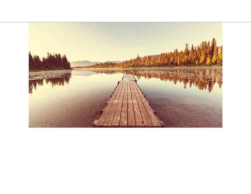 Ein langer Holzsteg führt über einen ruhigen See, umgeben von einem Wald und Bergen im Hintergrund, bei Sonnenuntergang.