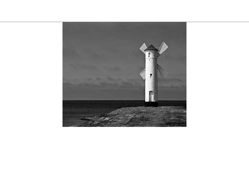 Ein alter Leuchtturm mit Windmühlenflügeln, stehend auf einem Steg am Meer, in Schwarz-Weiß aufgenommen.
